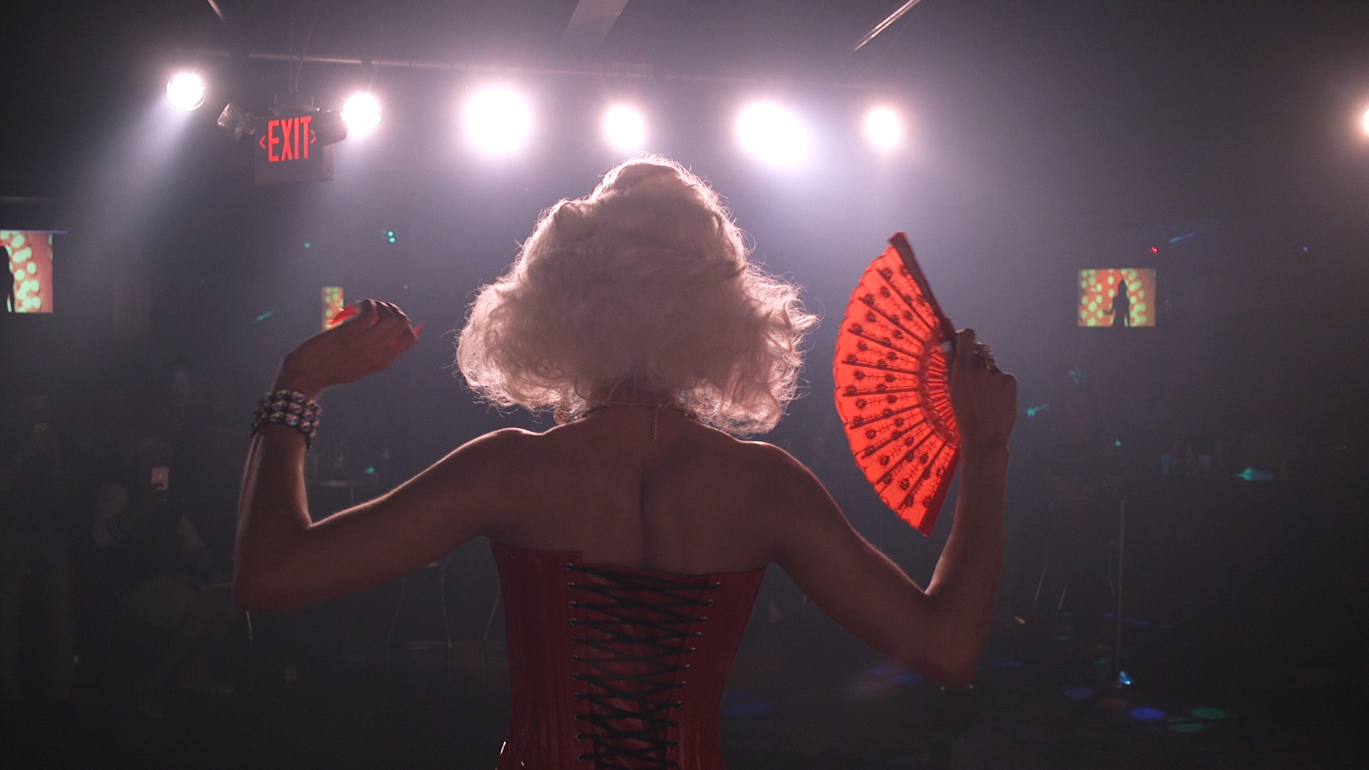 Cristina Morales performing in a blond wig, corset, and red fan, facing out into a dark bar and bright stage lights.