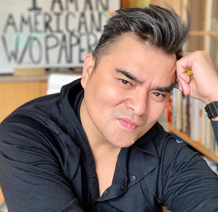 Headshot of Jose Antonio Vargas. In the background is a hand-lettered sign saying 'I am an American w/o papers.'