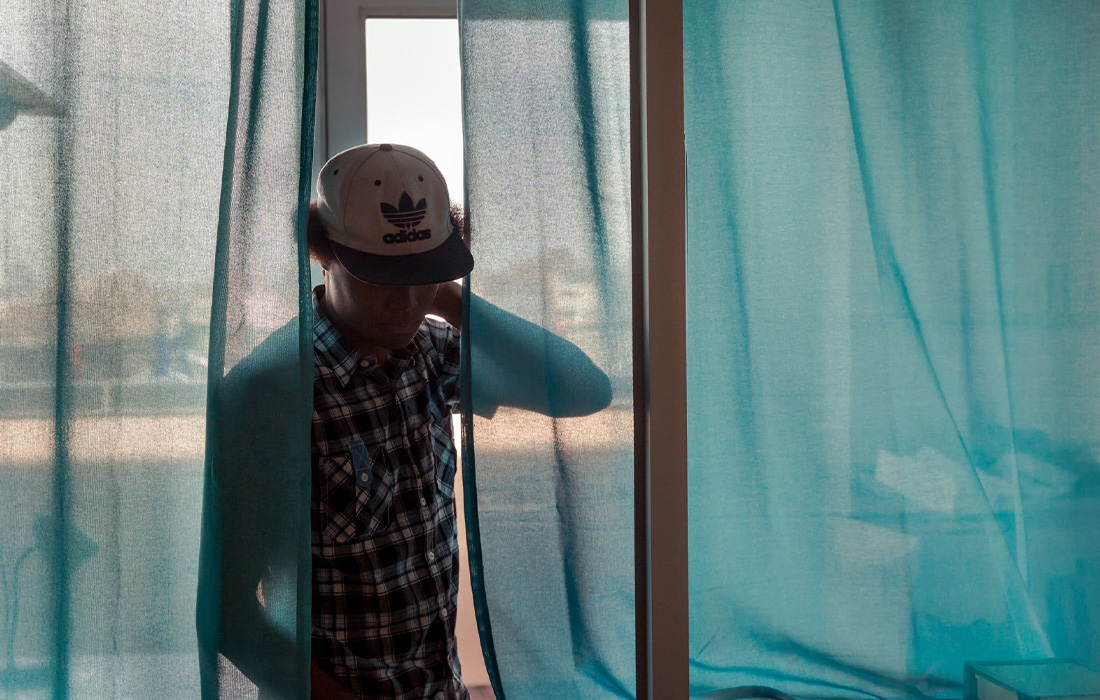 A man behind sheer blue curtains, face obscured by a baseball cap.